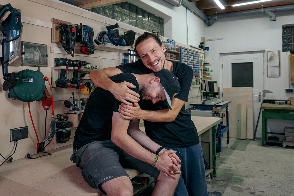 Zwei lächelnde Mitarbeiter von PARAX die schwarze T-Shirts tragen und sich in den Arm nehmen vor Werkzeugwand