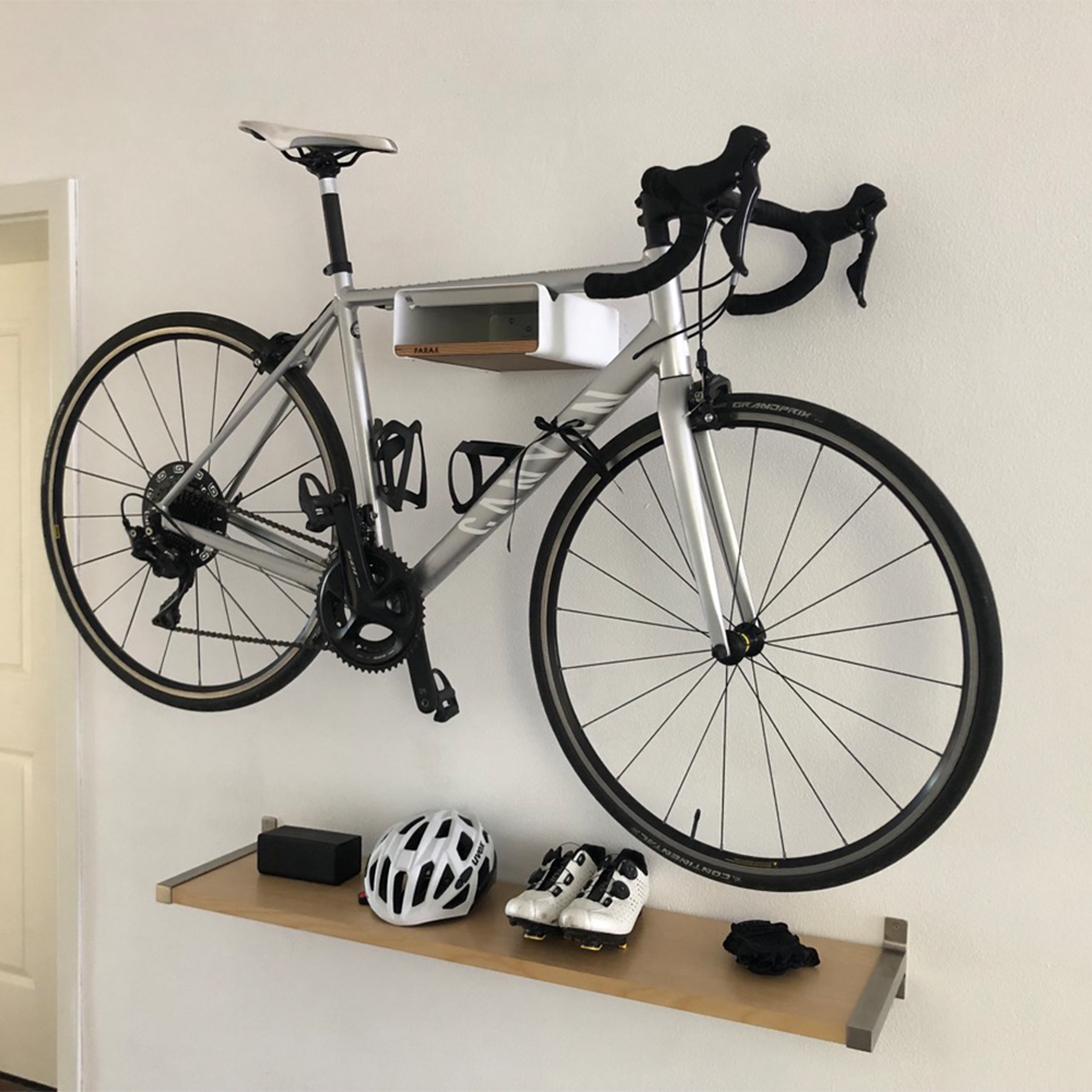 Weiße Fahrrad Wandhalterung S-RACK mit Einlegebrett und silbernem Fahrrad über Regal mit Fahrradhelm und Schuhen vor weißer Wand