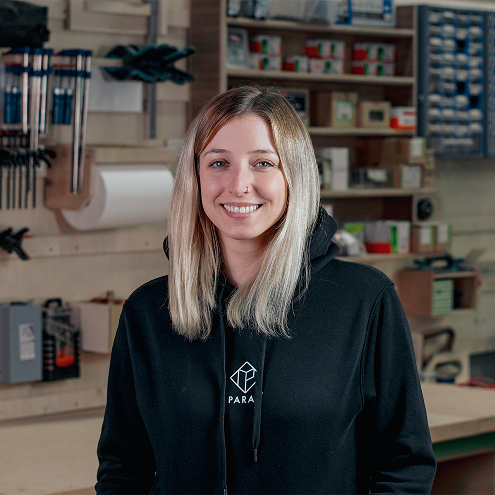 Lächelnde Mitarbeiterin von PARAX mit schwarzem Kapuzenpullover mit PARAX Logo vor einer Werkzeugwand
